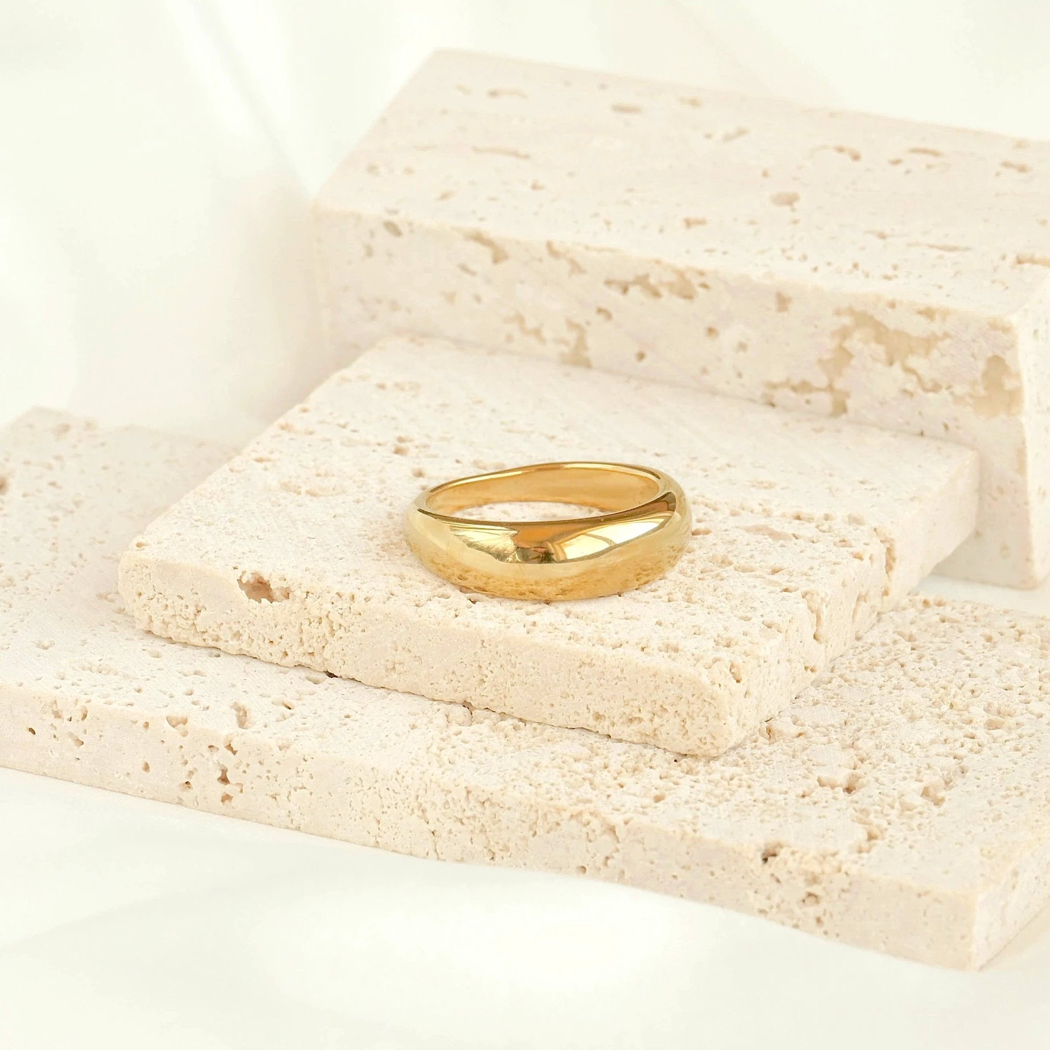 Shiny 18k gold dome ring with a thick, curved band resting on pale beige porous stone blocks.