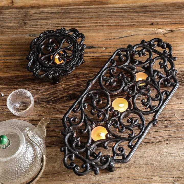 Ornate black cast-iron teapot warmer with lit tealights on a rustic wooden table beside a textured teapot.
