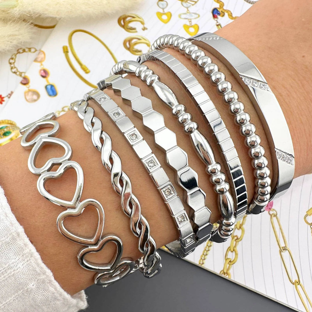Stack of silver bracelets on the wrist: heart links, twisted rope, hex links, beaded ball chain, rhinestone squares.
