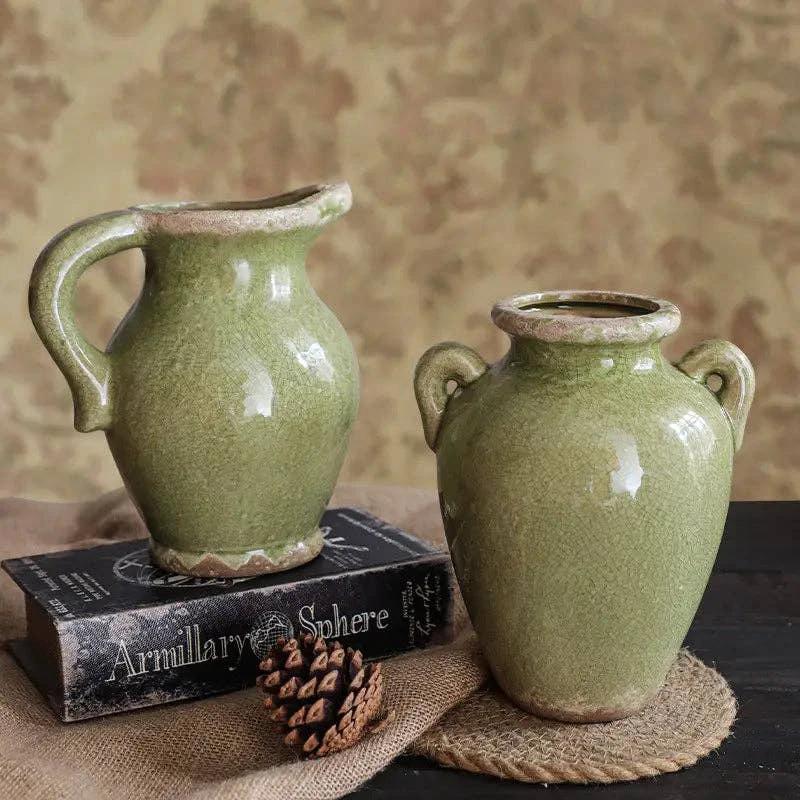 Olive-green glazed ceramic vases, a tall jug with curved handle and a twin-handled rounded vase, on a book with burlap.