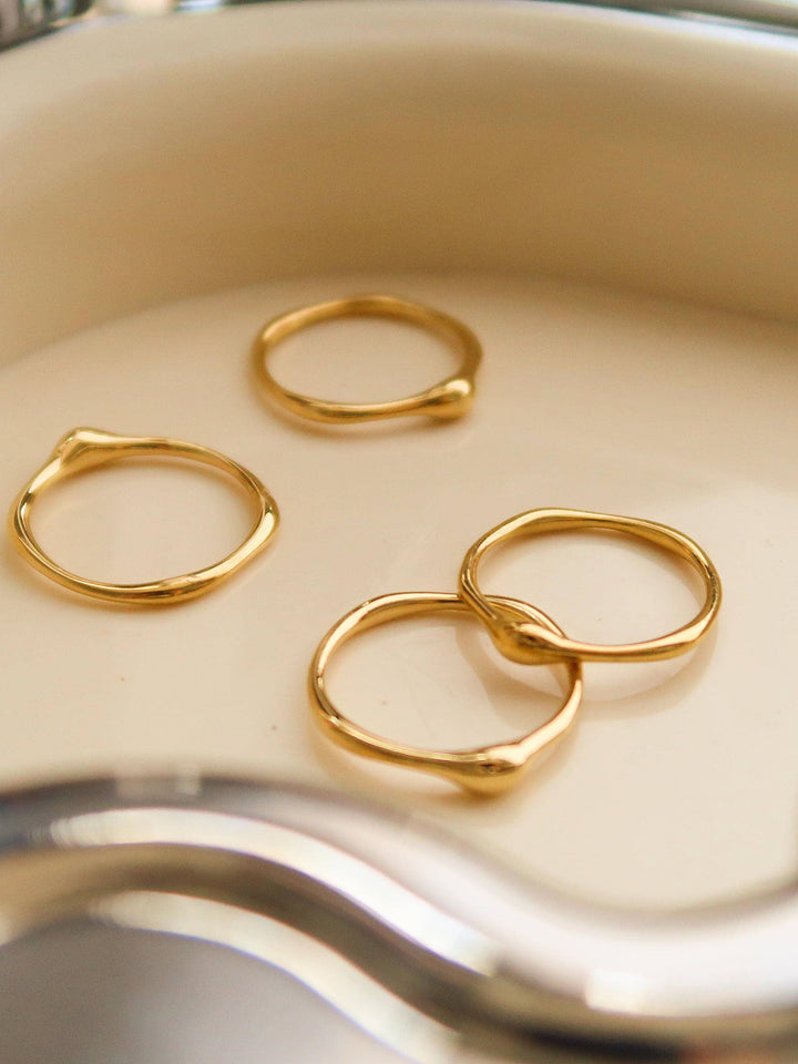 Four thin 18K gold minimalist band rings with organic, irregular shapes resting on a pale ceramic surface.