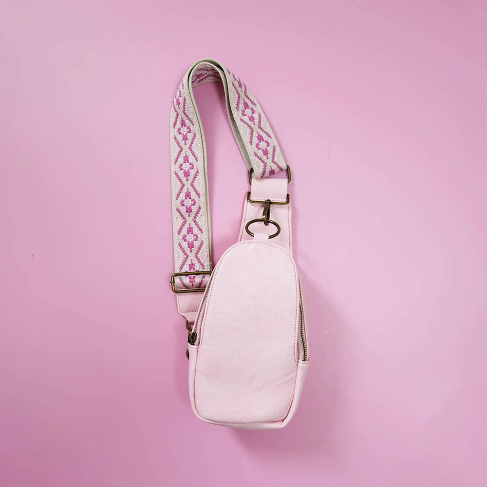 Pastel pink sling bag with a white-pink geometric guitar-style strap and brass hardware, against pink backdrop.