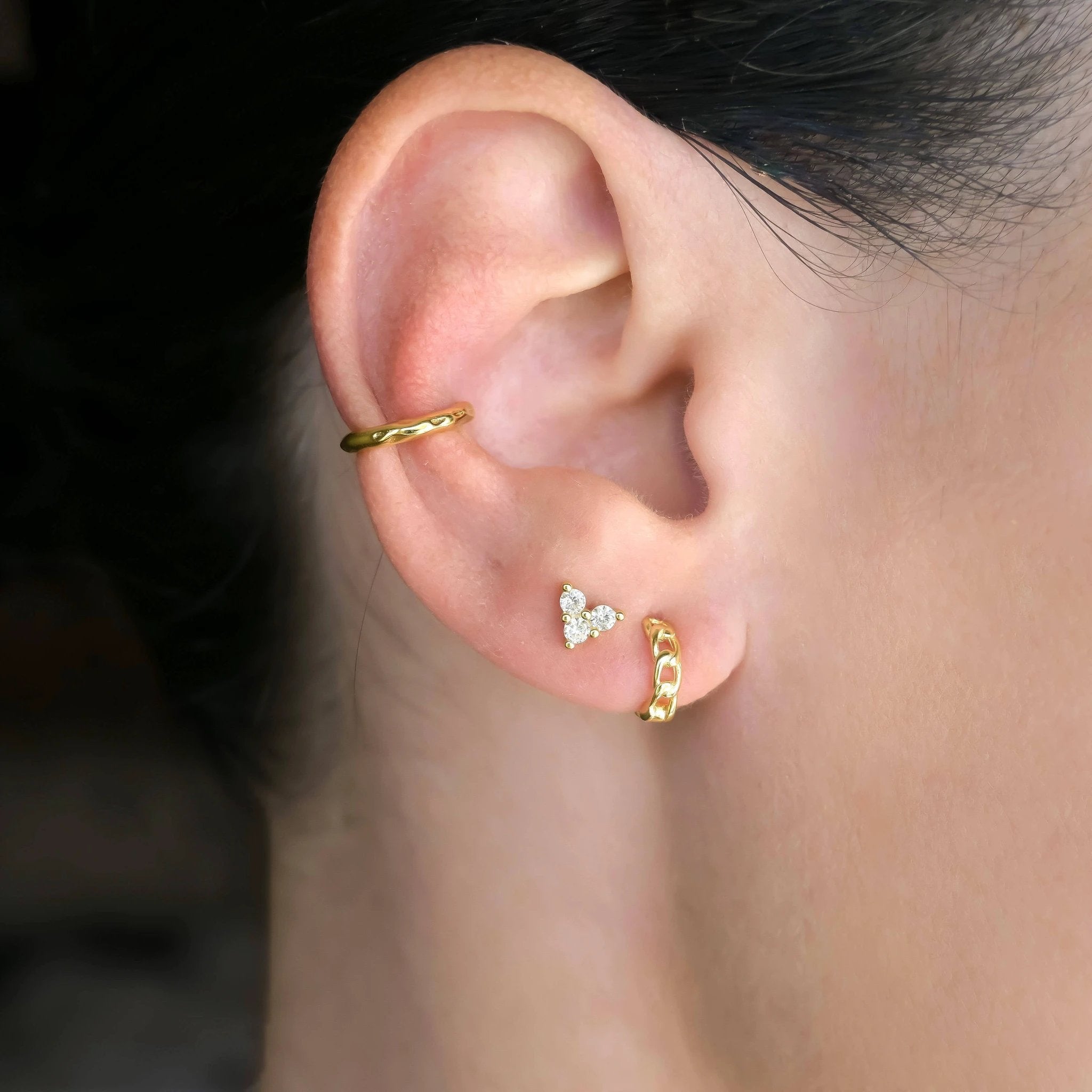 Gold-tone curved helix cuff, a diamond cluster stud, and a gold curb-link huggie hoop on the earlobe.