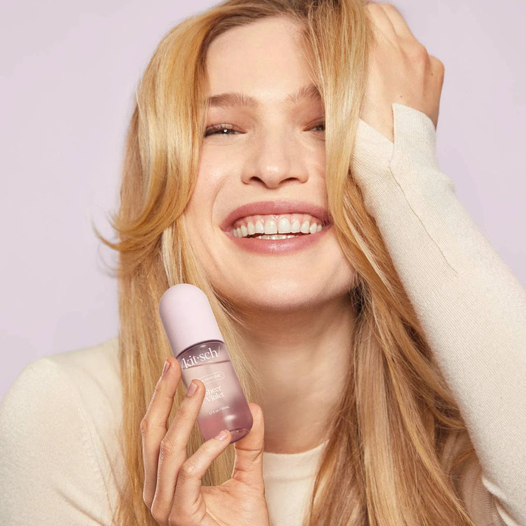 Sheer Violet Hair Perfume by KITSCH in a pink bottle held by a smiling blonde model