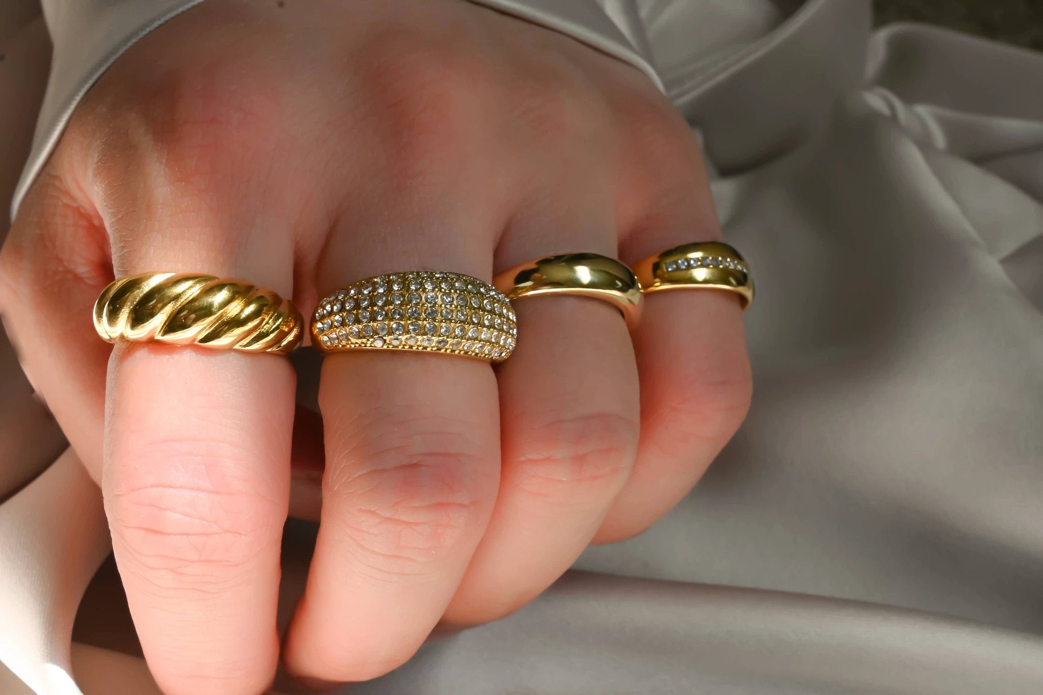 Close-up of four gold rings on fingers: twisted rope band, pavé-dome ring, thick smooth dome band, and slim band with stones.