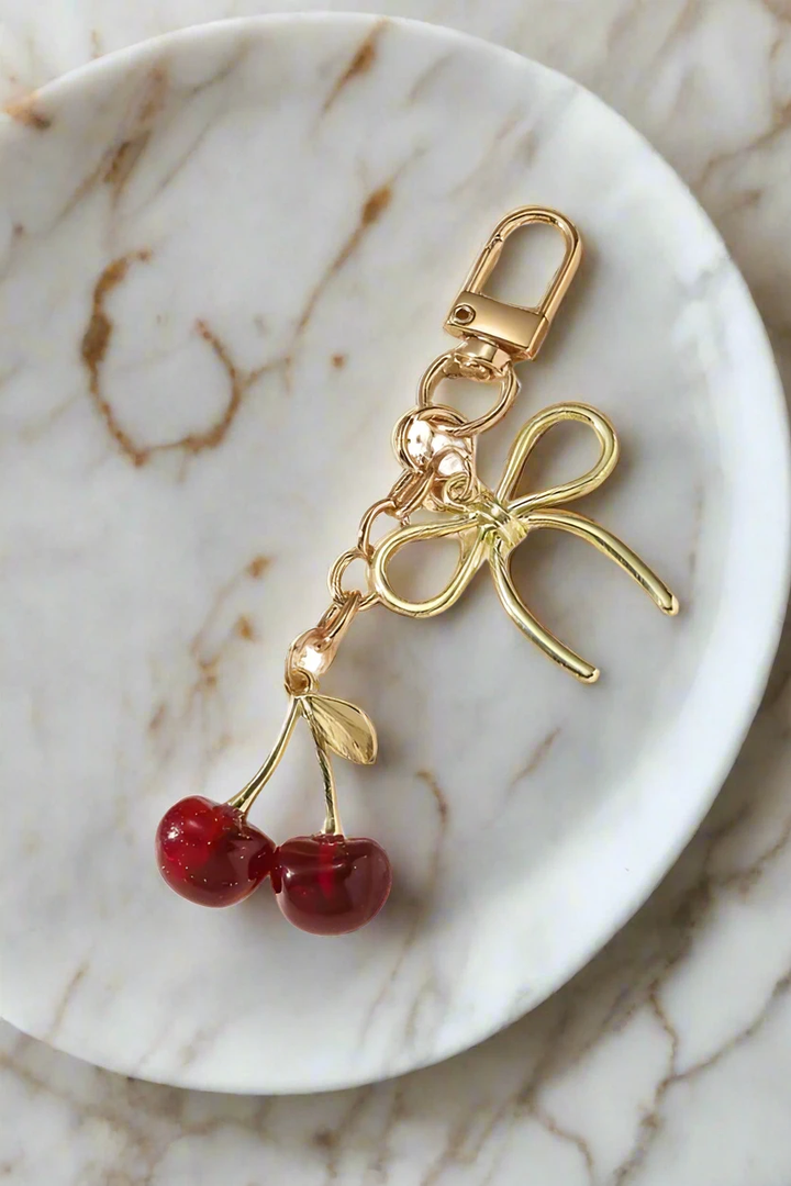 Gold-tone purse charm with bow, leaf, and two red cherry beads on a lobster clasp.