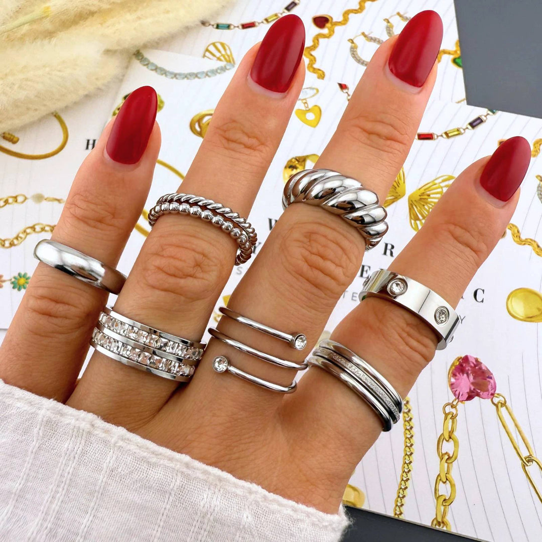 Hand with red nails wearing silver-tone stacking rings: twisted rope, beaded, and wide smooth bands with a small crystal accent.