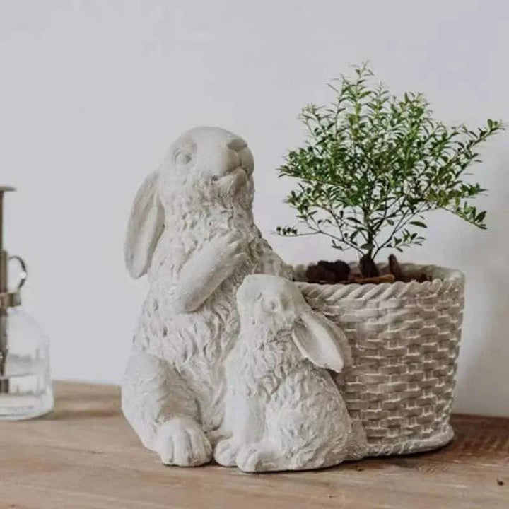 White cement double rabbit planter, with a small plant in a woven basket-style pot on a wooden surface.