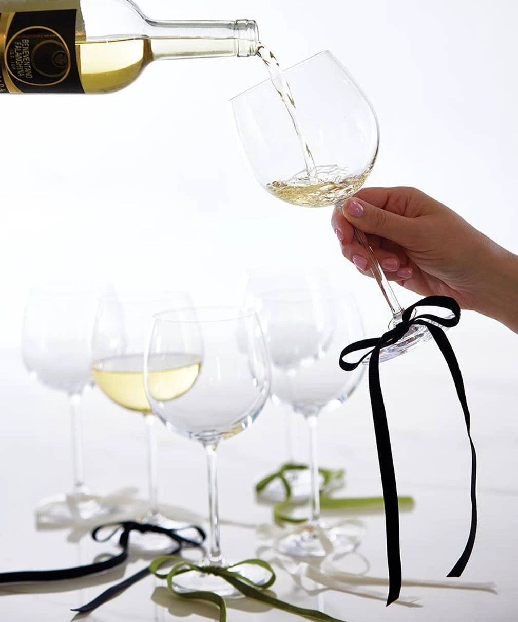 Six black velvet bow wine charms tied to wine glass stems, with a bottle pouring white wine into a glass in the background.