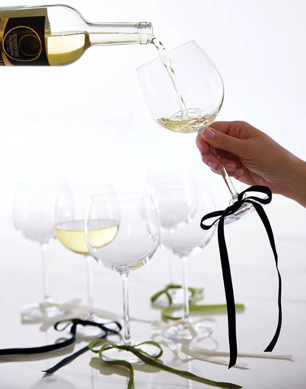 Six black velvet bow wine charms tied to wine glass stems, with a bottle pouring white wine into a glass in the background.