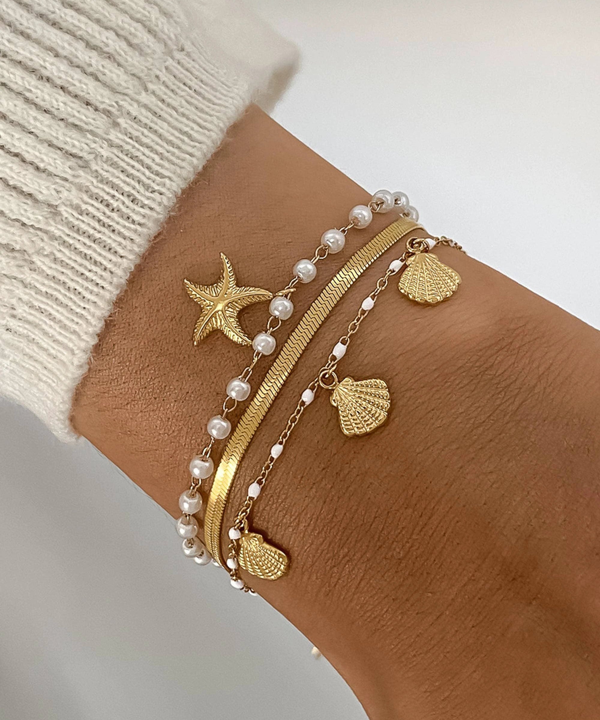 Gold-tone multi-layer bracelet stack with pearl beads, starfish charm, and shell charms on a knit wrist.
