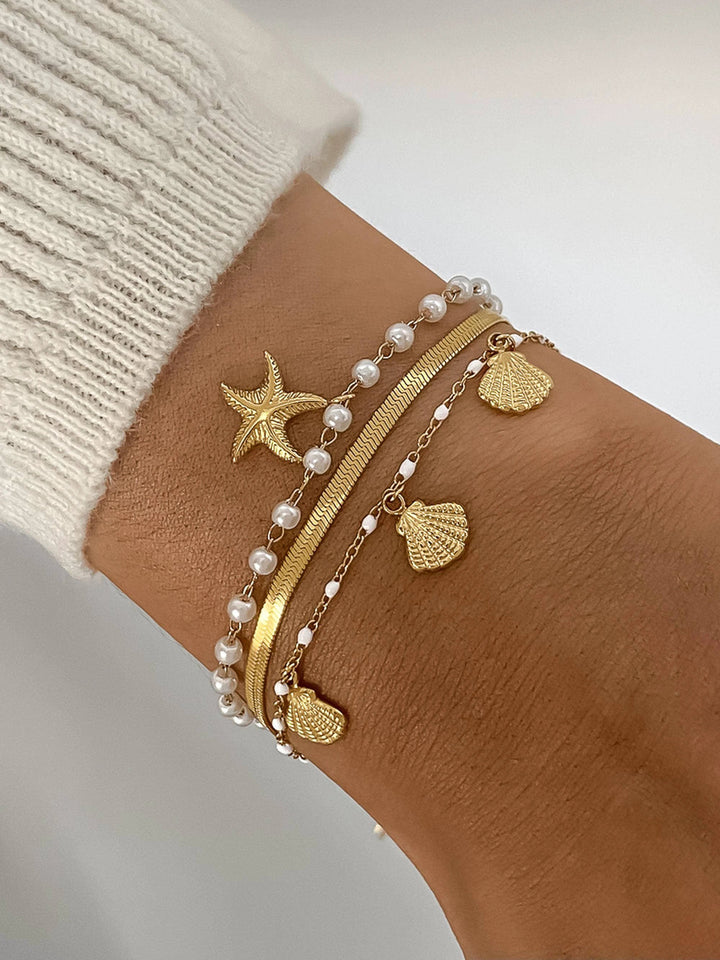 Gold-tone multi-layer bracelet stack with pearl beads, starfish charm, and shell charms on a knit wrist.