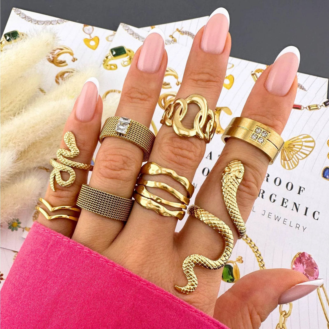 Hand with multiple gold-tone rings: twisted band, square-stone mesh band, chunky chain, and a snake wrap.