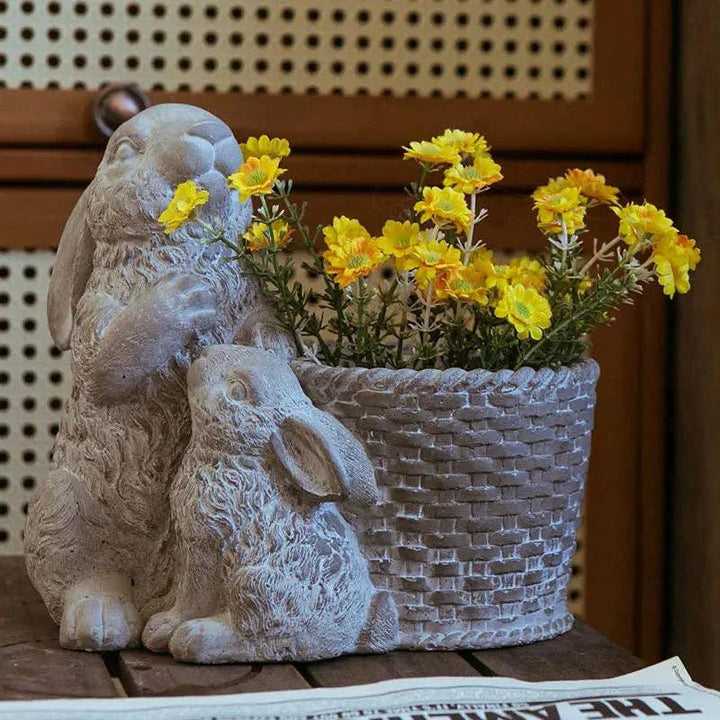 Two gray cement rabbits leaning against a brick-pattern planter filled with bright yellow flowers.