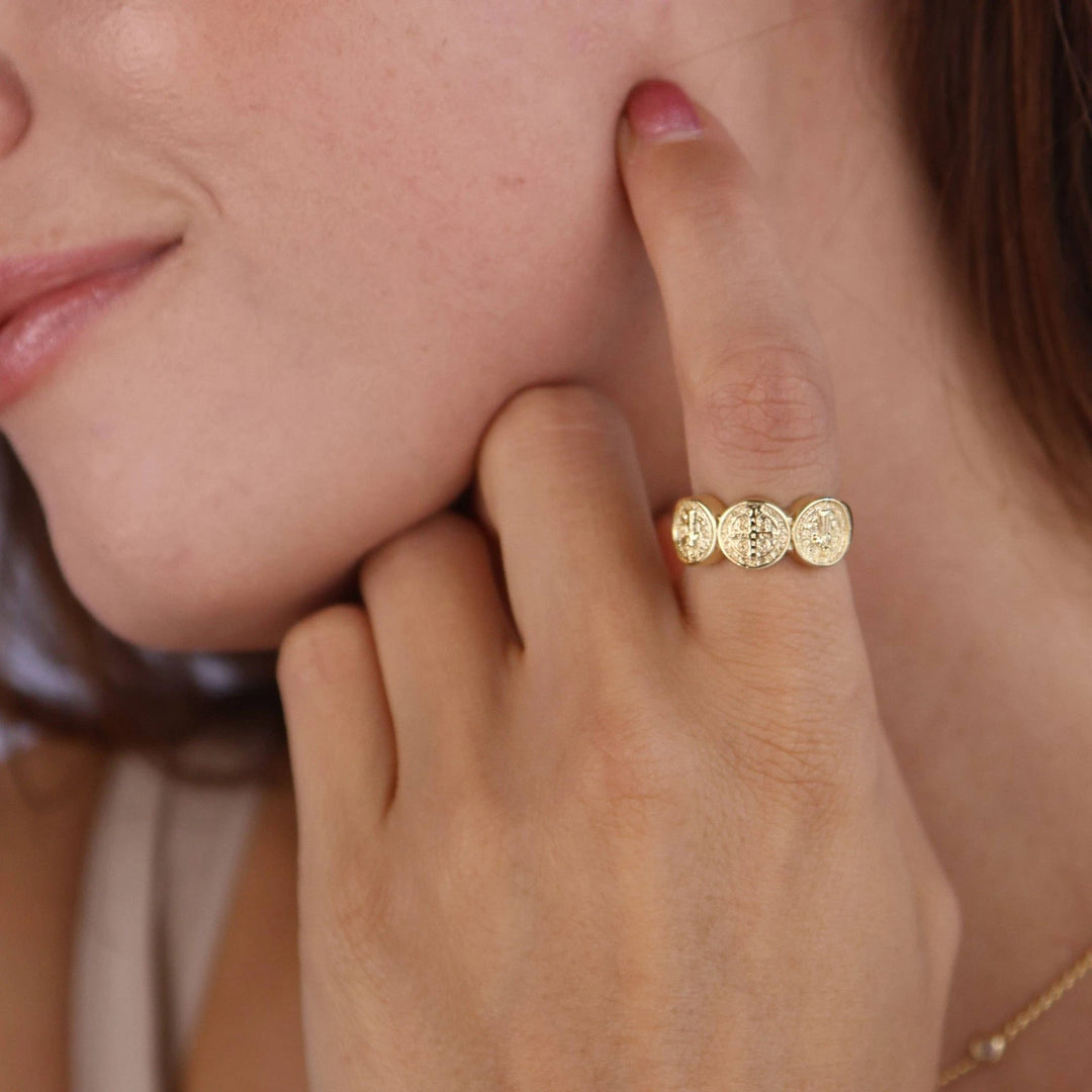 Close-up of a hand wearing an 18k gold-filled ring with three connected round Saint Benedict medallions.