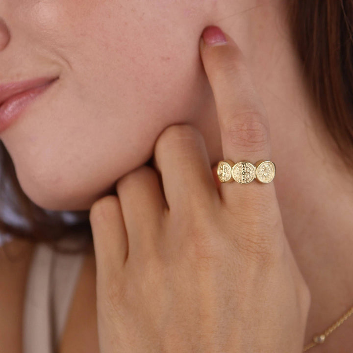 Close-up of a hand wearing an 18k gold-filled ring with three connected round Saint Benedict medallions.