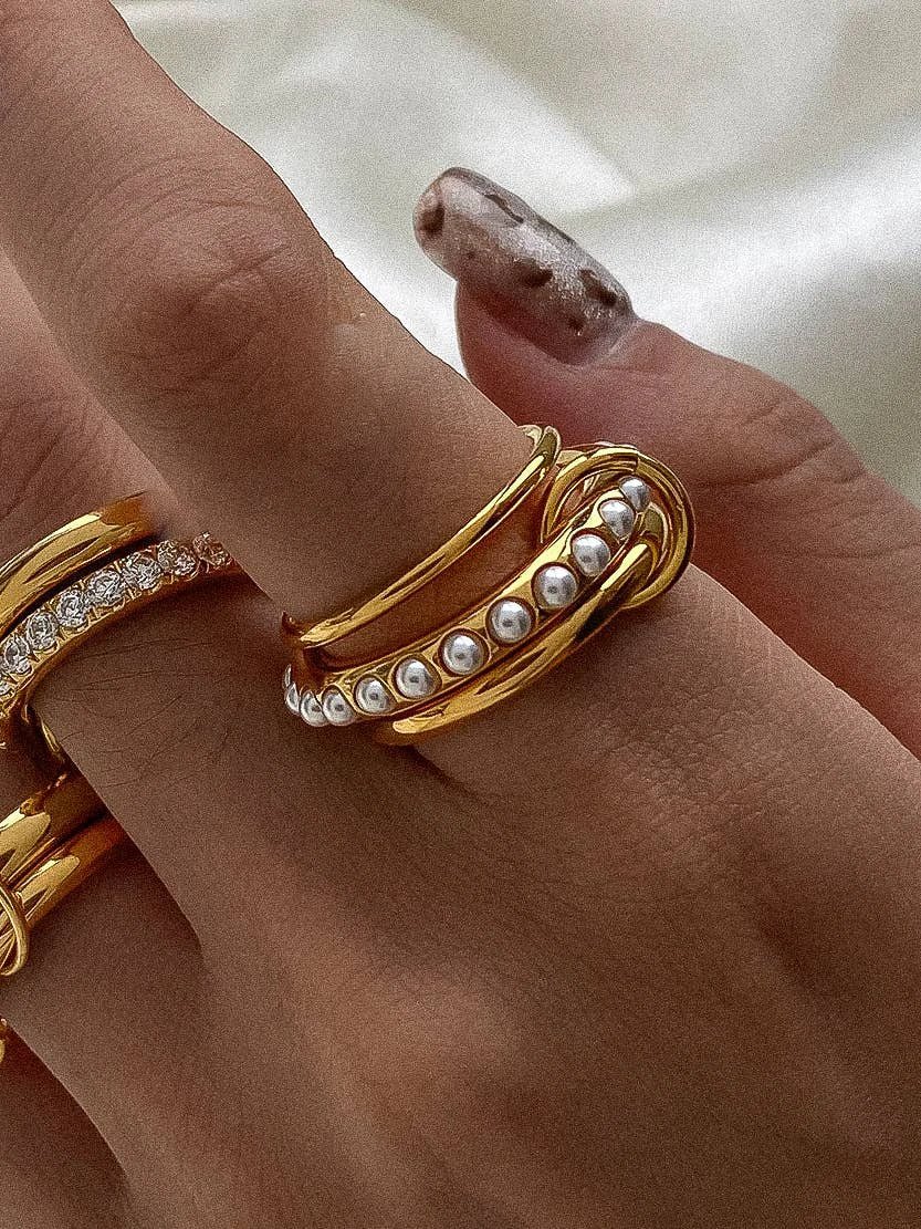 Stacked gold rings on finger: a diamond-studded band, a smooth gold band, and a pearl-embellished coiled ring.