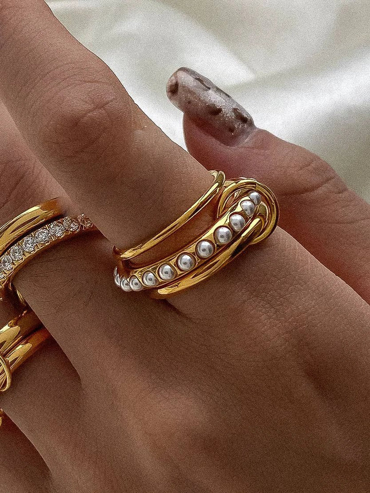 Stacked gold rings on finger: a diamond-studded band, a smooth gold band, and a pearl-embellished coiled ring.