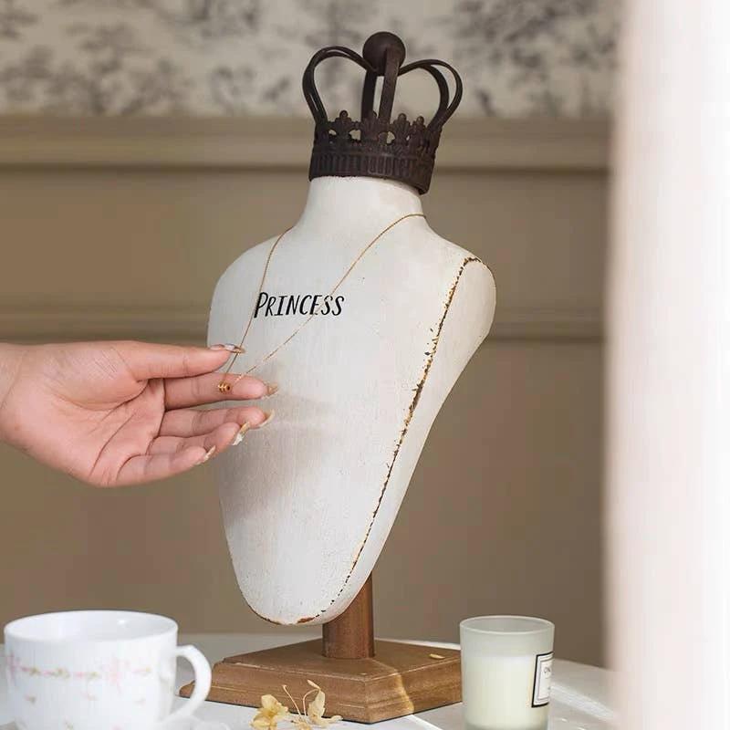 White resin neck form with a dark metal crown; a hand adjusts a gold chain across its chest near the word PRINCESS.