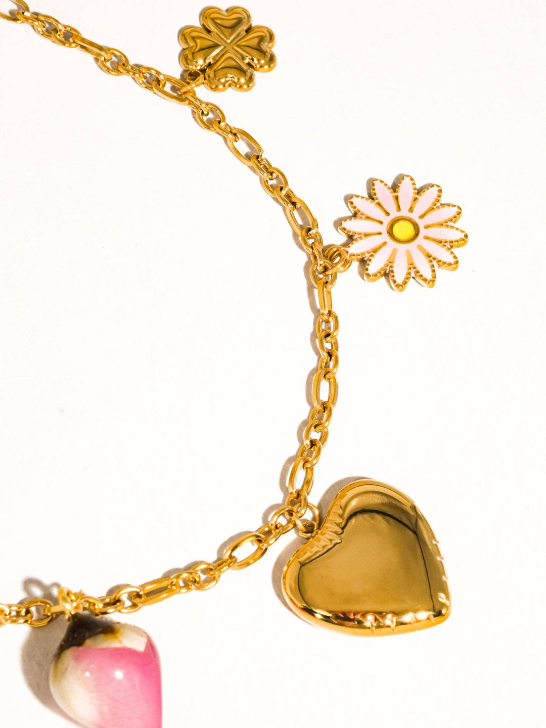 Gold chain necklace with clover charm, white enamel daisy, heart pendant, and pink teardrop bead.