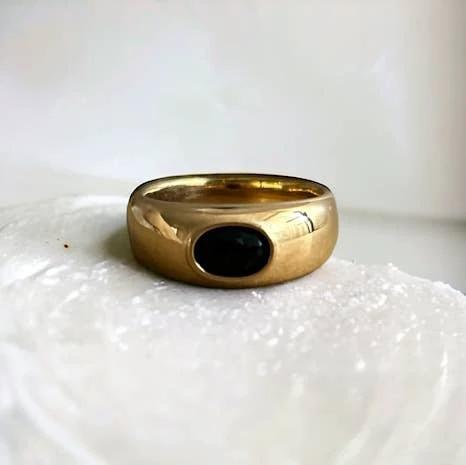 Gold-tone thick band ring with a black oval cabochon bezel-set stone, smooth polished finish resting on a white textured surface.
