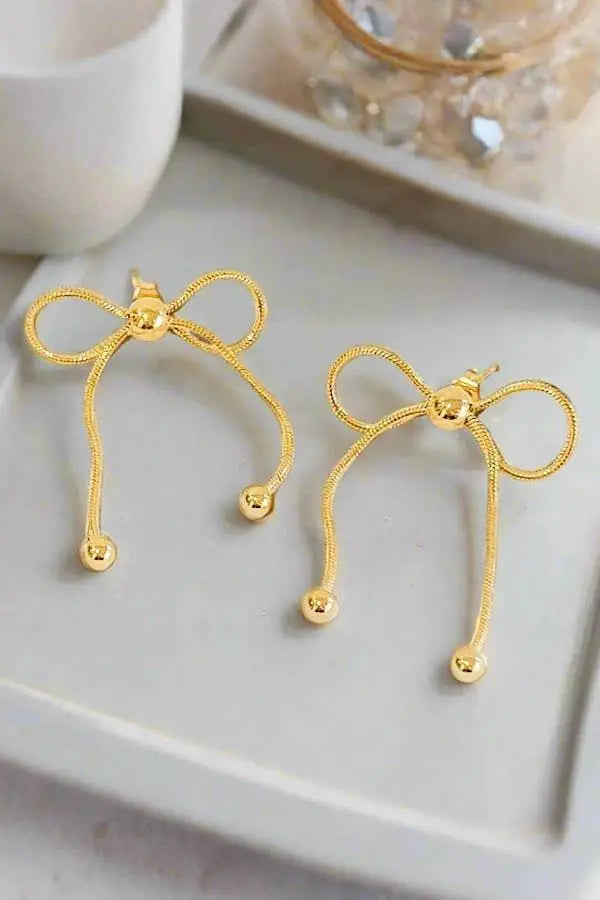 Gold-tone bow-shaped dangle earrings with looped bows and snake-chain tails ending in small gold beads, displayed on a light gray tray.