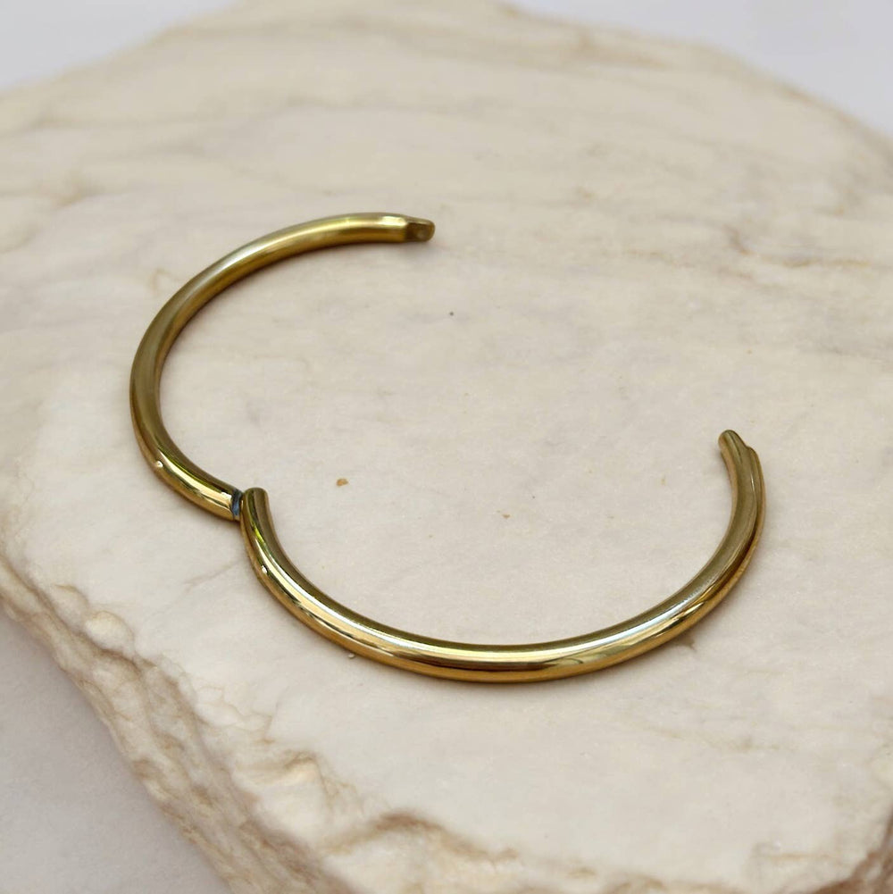 Gold-tone open cuff bangle with a smooth curved tube, resting on a light stone surface.