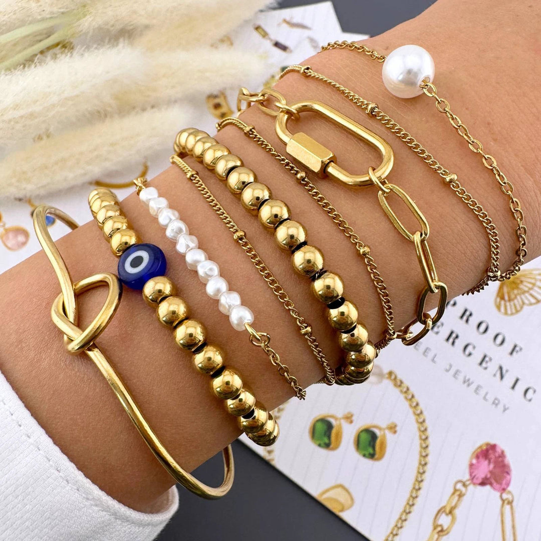 Layered gold-plated stainless steel bracelets with pearls, white beads, a blue eye charm, and delicate chain links on a wrist.