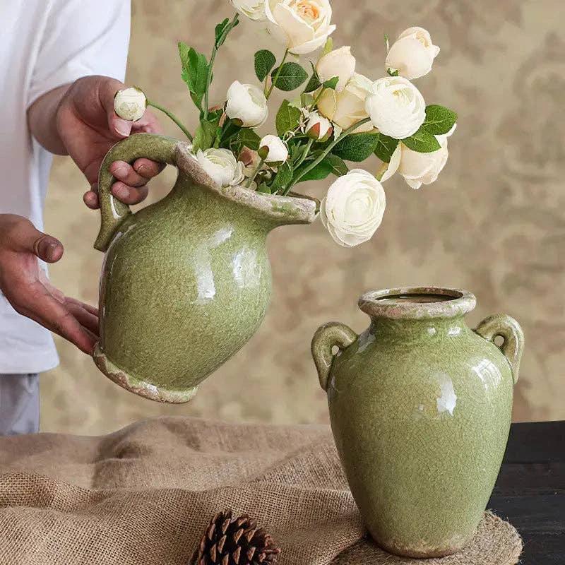 Two olive-green glazed ceramic vases with curved handles; one held by hands with white roses, the other on burlap.