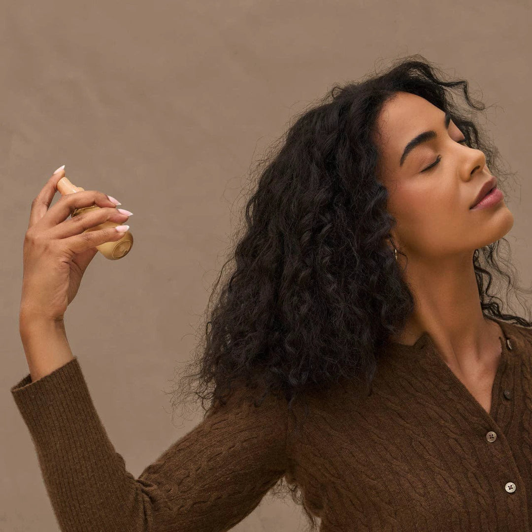 Rustic Vanilla Hair Perfume bottle in beige, held by a woman with curly dark hair wearing a brown knit top.
