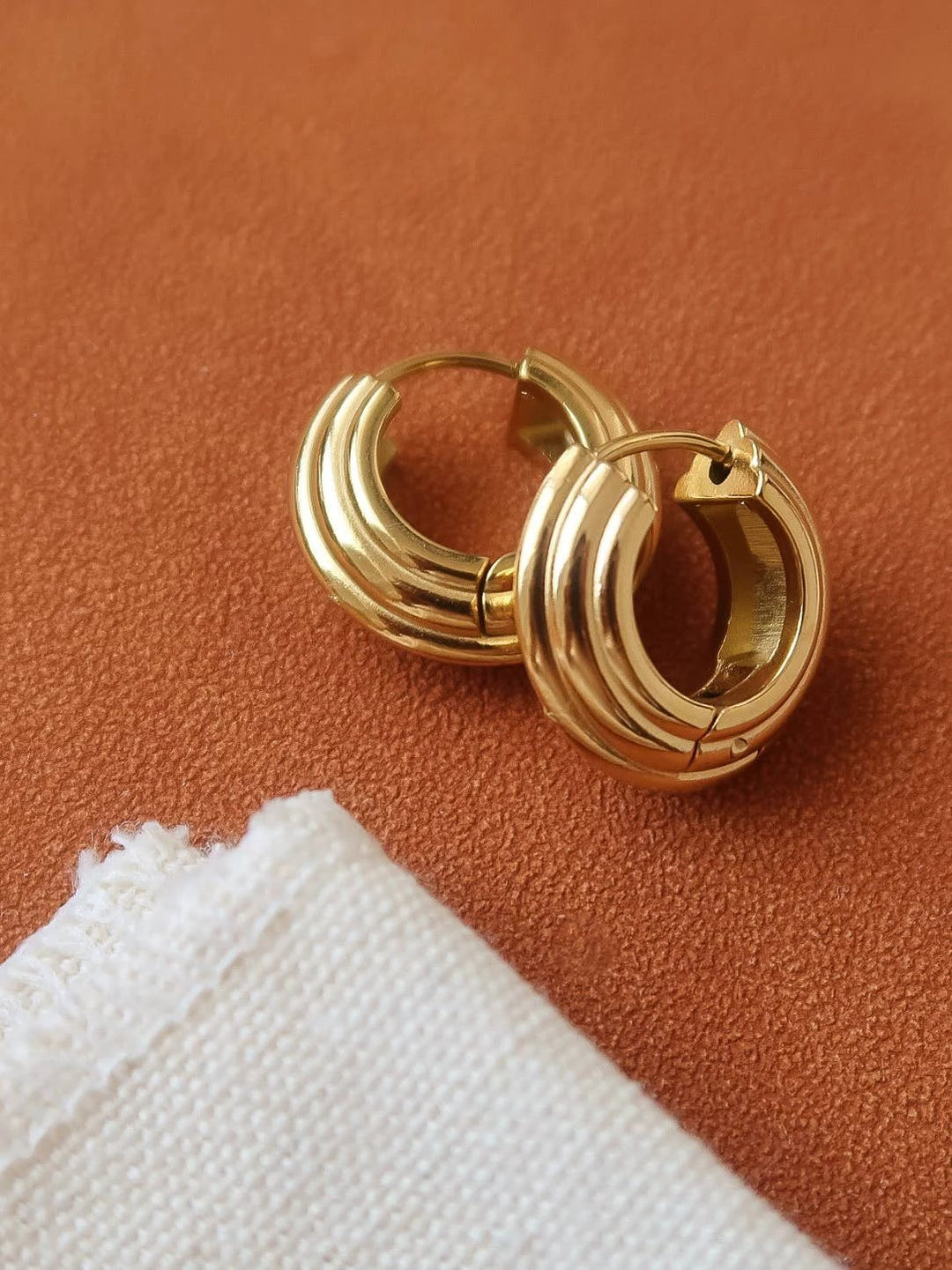 Two thick gold-tone hoop earrings with triple-rim design and hinged clasps, resting on a brown textured surface with a white fabric corner.