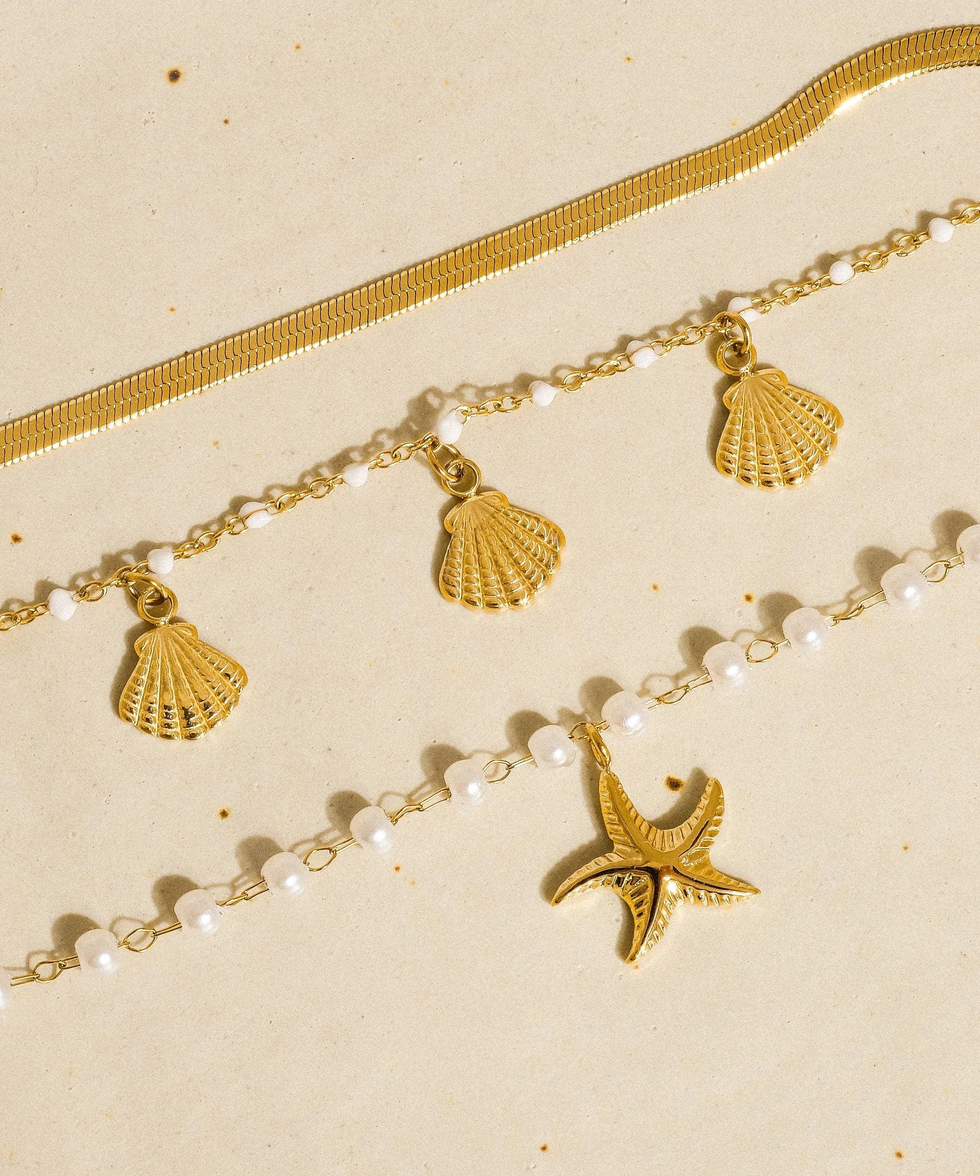 Layered 18K gold bracelet set with shell charms, white pearl beads, and a starfish charm on a beige background.