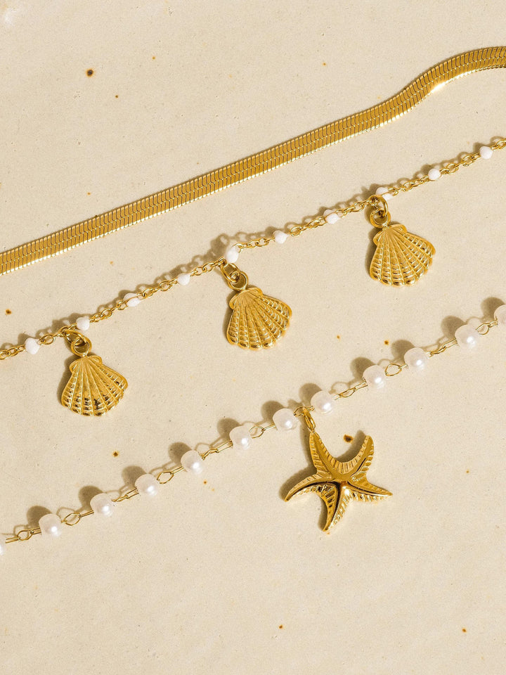 Layered 18K gold bracelet set with shell charms, white pearl beads, and a starfish charm on a beige background.