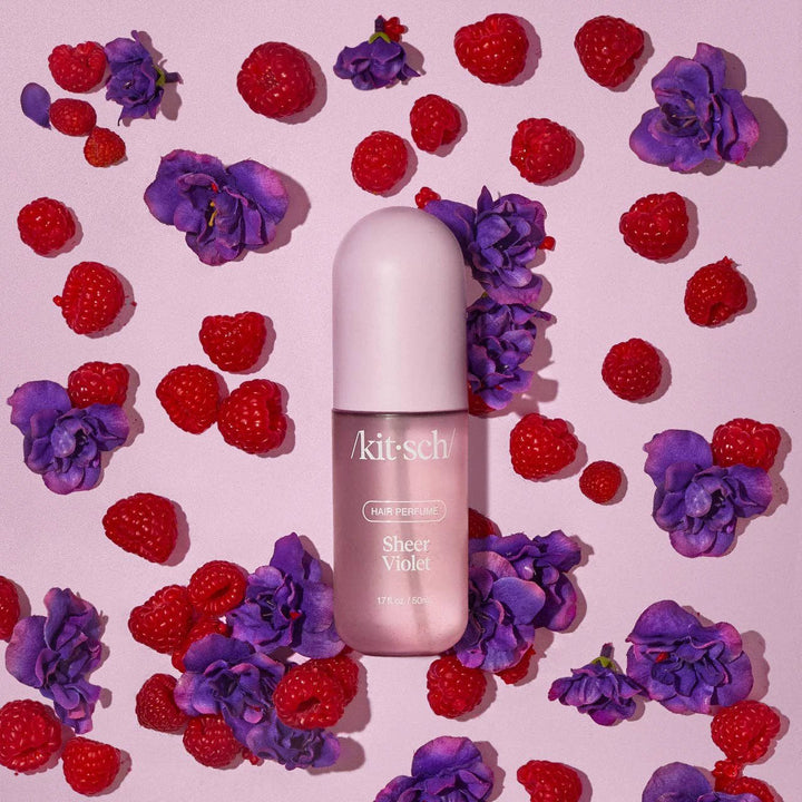 Kitsch Sheer Violet Hair Perfume bottle in pink, surrounded by purple flowers and raspberries