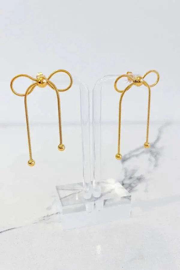 Pair of gold-tone bow-dangle earrings with long hanging chains ending in small beads, on a clear display stand.