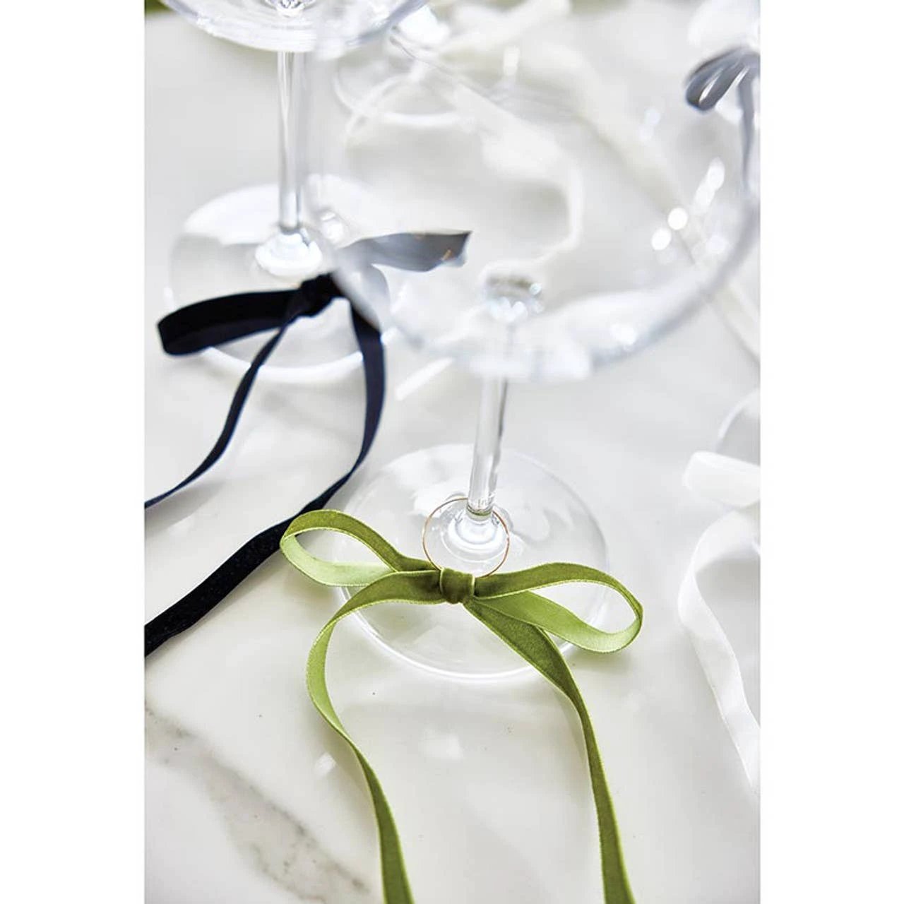 Green velvet bow wine charm tied around a wine glass stem with a gold ring detail; black ribbon in the background.
