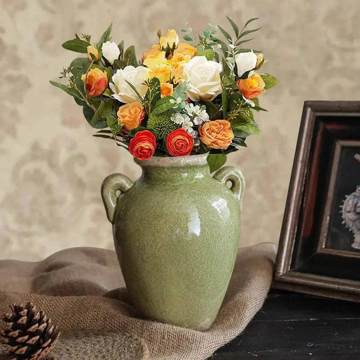 Green glazed ceramic vase with two handles, orange, yellow, and white roses with greenery on burlap, pine cone nearby.