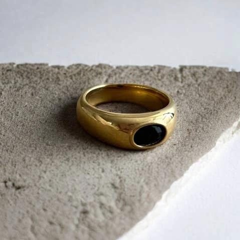 Gold signet ring with a black oval bezel-set stone, resting on a textured gray stone surface.