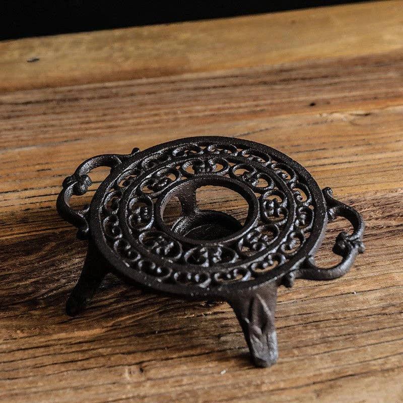 Black cast-iron teapot warmer with ornate circular scrollwork, curved side handles, and three legs on a wooden surface.