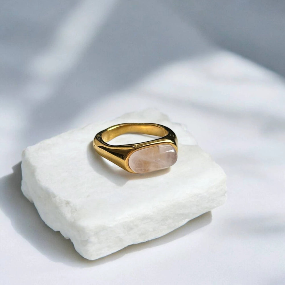 Bold gold ring featuring a pale pink oval cabochon stone set in a smooth bezel, resting on a white stone block.