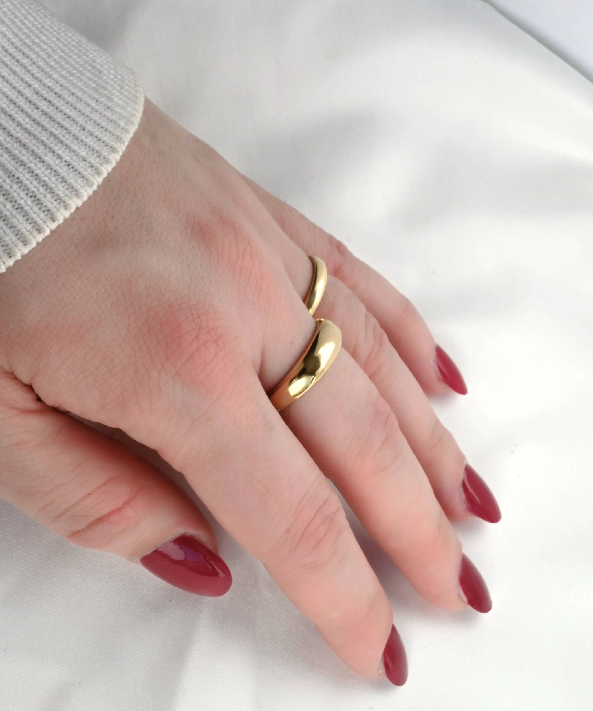 Close-up of a hand wearing two glossy 18k yellow gold dome rings stacked on a finger, red nails and a light knit cuff.