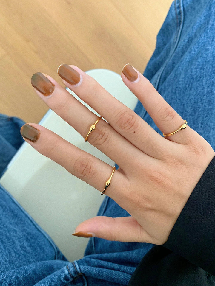 Hand with brown manicure wearing several thin 18K gold curved-band rings; minimalist style, denim backdrop.