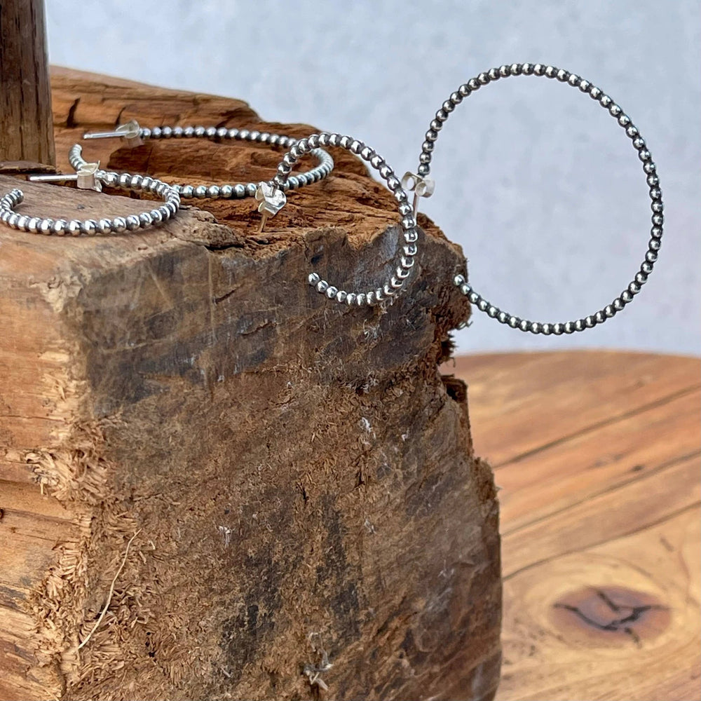 Sterling silver beaded hoop earrings laid on a rough wooden block; tiny beads form curved hoops with clear earring backs.