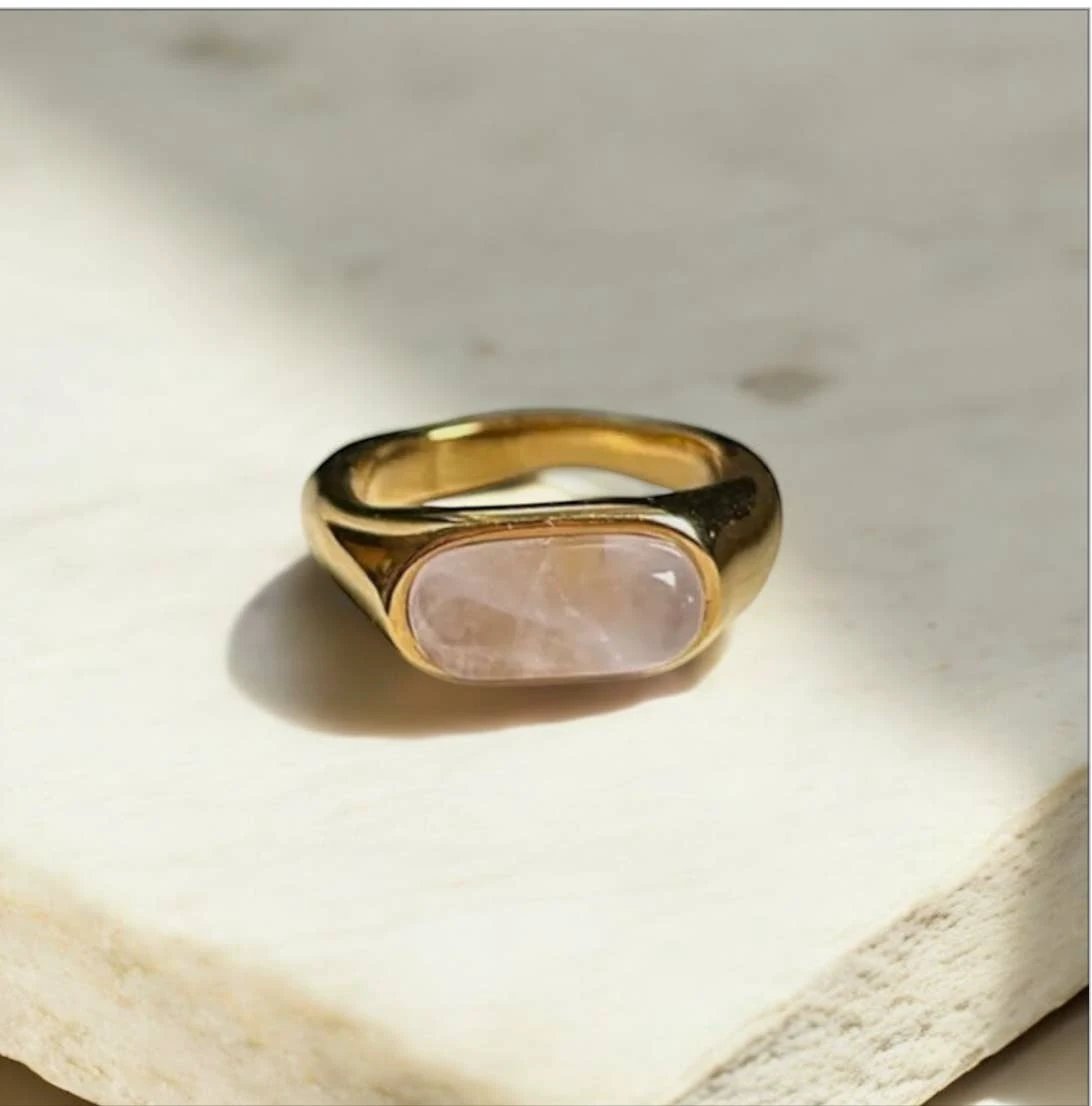 Amara Ring in gold with a wide bezel-set pale pink rectangular cabochon, resting on a light marble surface.