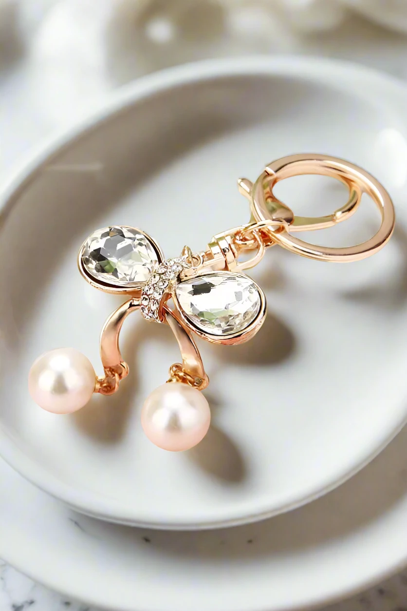 Rose-gold purse charm with a crystal bow, two clear oval stones, rhinestone center, and two pink pearl drops on a ring.