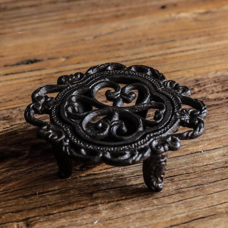 Ornate black cast-iron teapot warmer with circular scrollwork, raised rim, and three short legs on a wooden surface.
