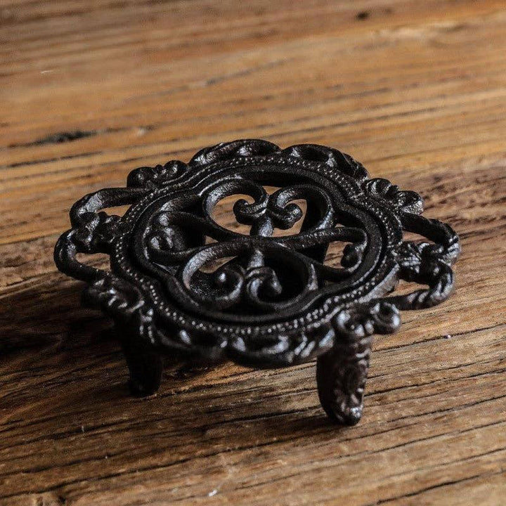 Ornate black cast-iron teapot warmer with circular scrollwork, raised rim, and three short legs on a wooden surface.