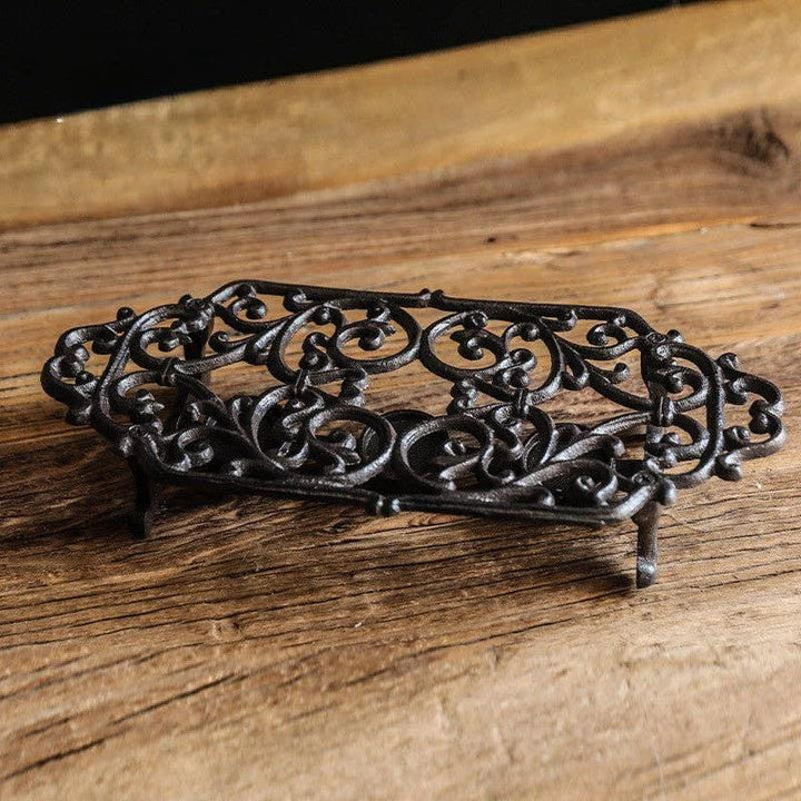 Ornate black cast-iron teapot warmer stand with intricate scrollwork, raised on small feet on a wooden surface.