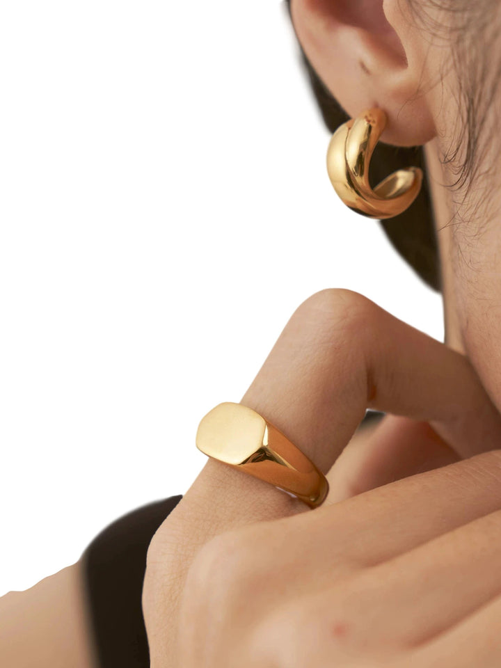 Polished 18k gold hexagon signet ring on finger; chunky curved hoop earring visible in ear.